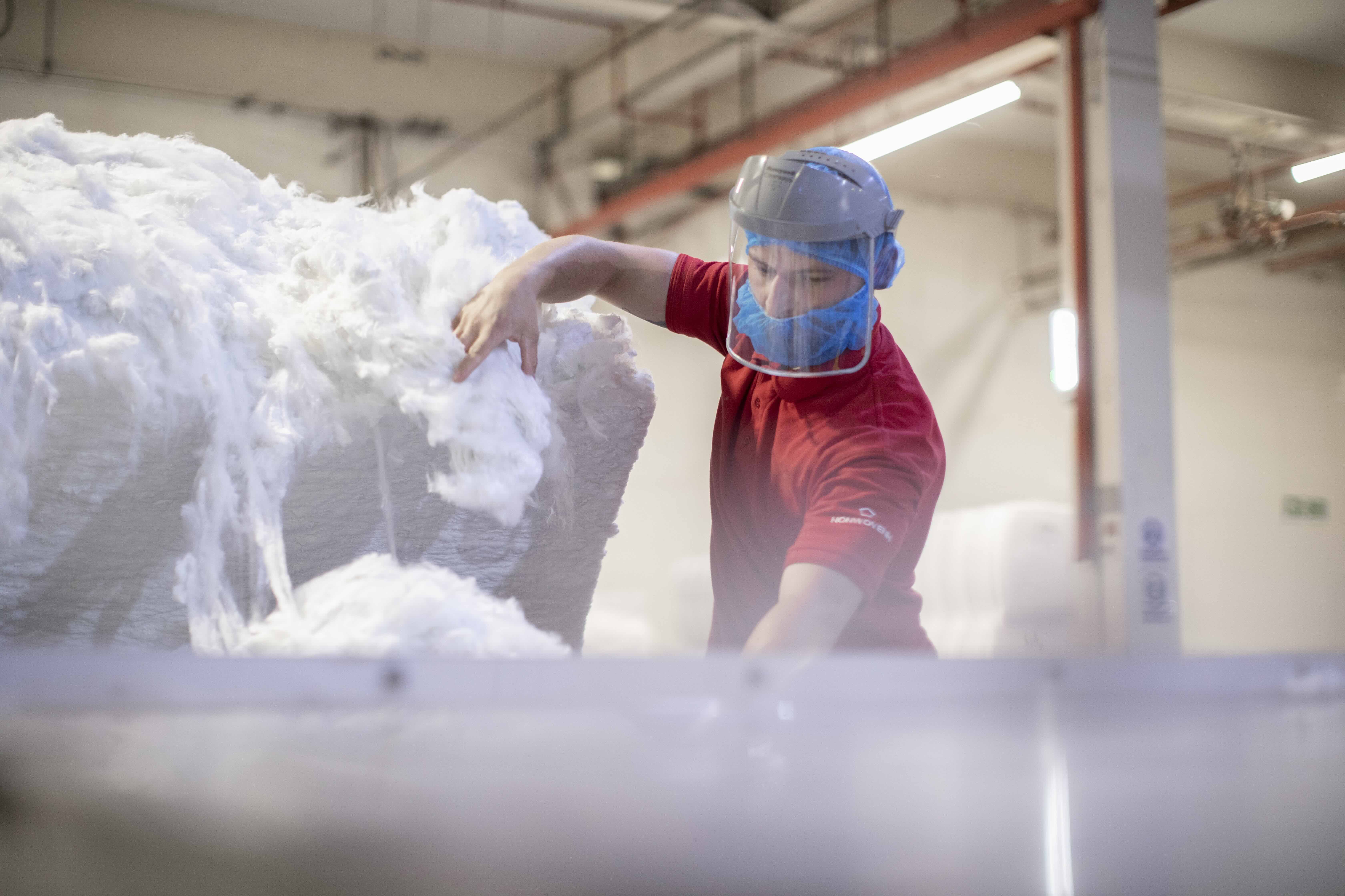 A worker in a red shirt and blue hairnet handles fluffy white material in a bright industrial setting, showcasing nonwoven fabric production.
